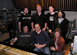 Seven men smiling together in a music studio with recording equipment and a mixing console.