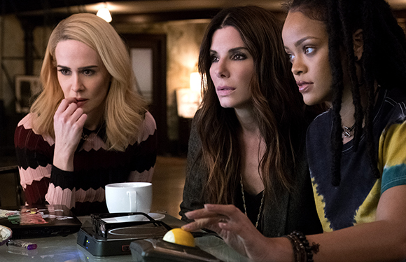 Three women with focused expressions lean forward toward a table in a dimly lit room.