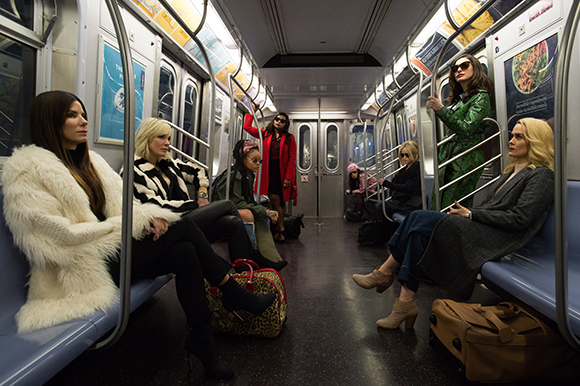 Six women in stylish clothing sit on blue subway seats in a brightly lit train car.