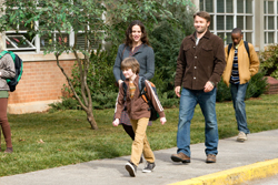 A man, woman, and child walk along a sidewalk past a brick building and green bushes.
