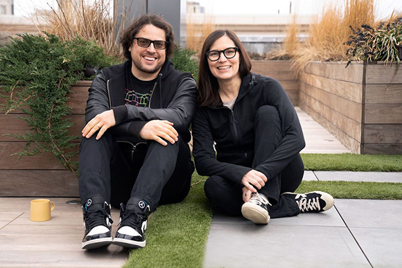 A man and a woman in black clothing sit on a rooftop patio next to planters and grass.