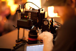 A person's hands adjusting a professional RED cinema camera mounted on a rig in a dimly lit studio.