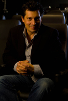 A man in a dark blazer and light shirt sits with hands clasped in a dimly lit theater.