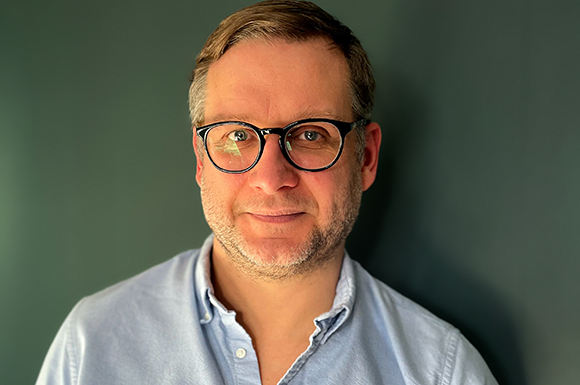 A man with short brown hair and glasses wearing a light blue button-down shirt against a dark green background.