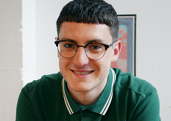 A young man with short dark hair and glasses smiling while wearing a green polo shirt.
