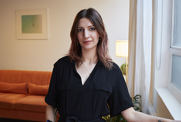 A woman with long brown hair wearing a black short-sleeved shirt stands in a bright room.