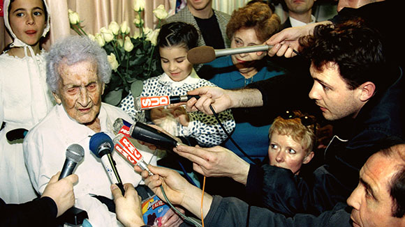 An elderly woman is surrounded by several people holding microphones toward her.
