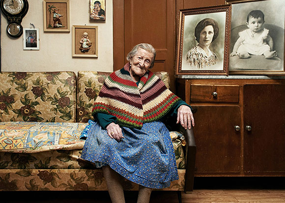 An elderly woman wearing a colorful crocheted shawl sits on a floral patterned sofa.