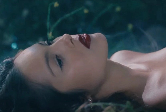 Close-up profile of a person with dark hair and red lipstick lying down against a dark, blurry background.