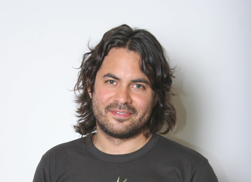A man with wavy dark hair and a light beard smiling against a white background.