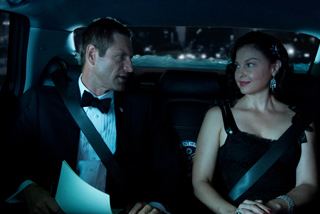 A man in a tuxedo and a woman in a black dress sit in the backseat of a car at night.