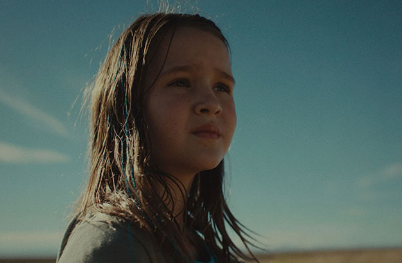 A young girl with long brown hair looks thoughtfully toward the horizon under a clear blue sky.