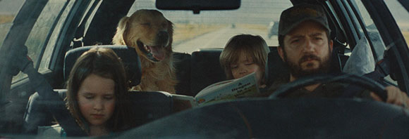 A man driving a car with two children and a golden retriever in the backseat.