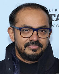 A man with dark hair, glasses, and a beard wearing a dark coat.