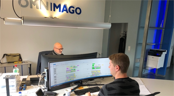 Two men sit at desks with large computer monitors in an office with an OMNIMAGO sign on the wall.