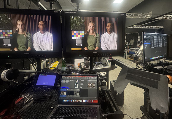 Multiple computer monitors displaying video footage of two people and editing software in a dark studio.