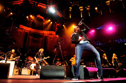 A male singer in a black jacket and jeans performs with a microphone on a brightly lit concert stage.