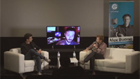Two men sit in white armchairs facing a large television screen in a dark room.