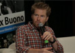 A man with reddish hair wearing a plaid shirt speaks into a handheld microphone.
