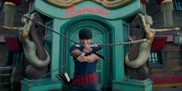 A man in a black cap holds two long swords crossed in front of a green building named Baratie.
