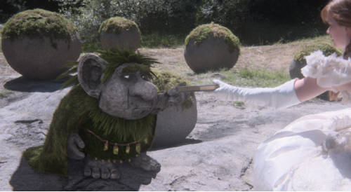 A woman in white gloves reaches toward a small, mossy stone troll statue outdoors.