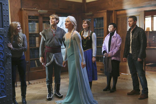 Six people in various costumes stand in a library-like room with wood paneling and bookshelves.