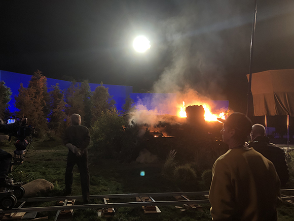 Film crew watching a large fire and smoke on a dark outdoor set under a bright moon.