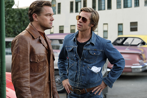 A man in a brown leather jacket looks at a man in a denim jacket and sunglasses.
