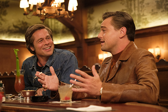 Two men sit at a wooden bar, one in a denim jacket and one in a brown leather jacket, gesturing while talking.