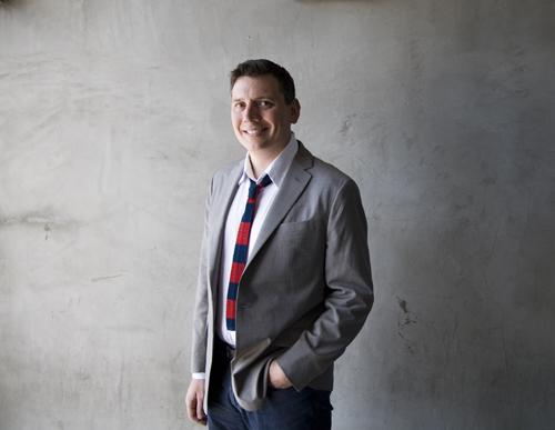A smiling man wearing a grey blazer, white shirt, and red patterned tie stands against a grey wall.