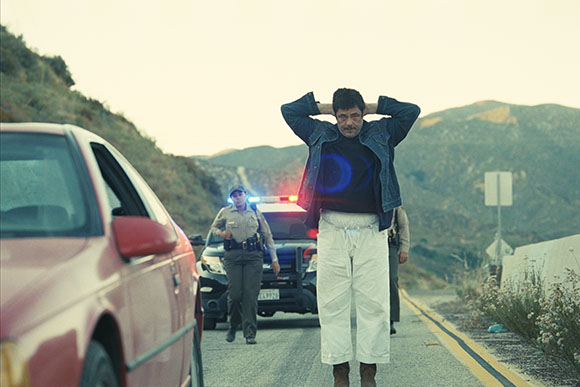 A man stands with hands behind his head on a road near a red car and a police vehicle.