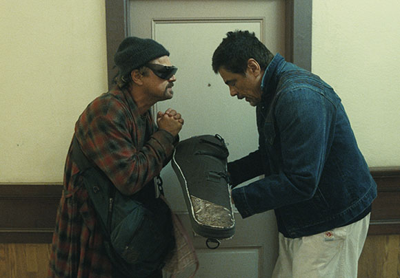 Two men stand facing each other in a room, one holding a dark bag toward the other.