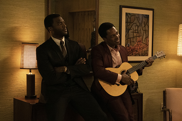 Two men in suits pose in a room, one holding an acoustic guitar.