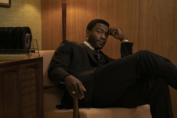 A man in a black suit and tie sits in a chair, resting his head on his hand with a serious expression.