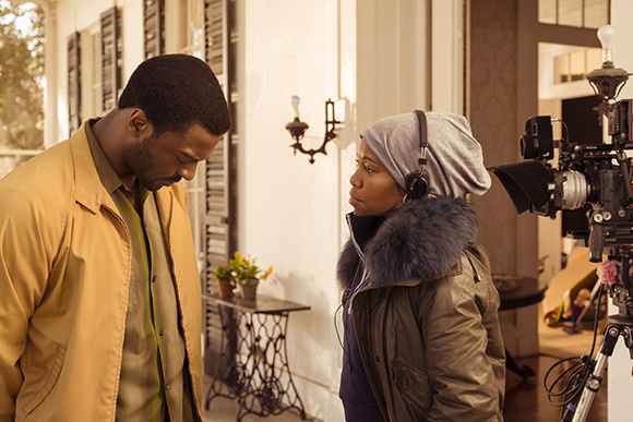 A man in a tan jacket looks down while a woman in a grey hood and headset looks at him during a film shoot.