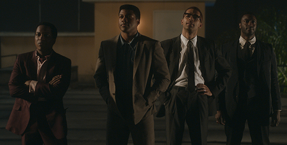 Four men in formal suits stand together outdoors under dim lighting at night.