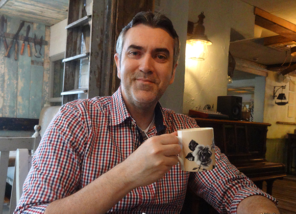 A man in a red and white checkered shirt holds a white mug with a floral design in a dimly lit room.