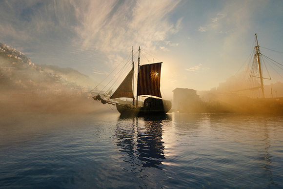 A wooden sailing ship with striped sails floats on calm water amidst morning mist near a coastal town.