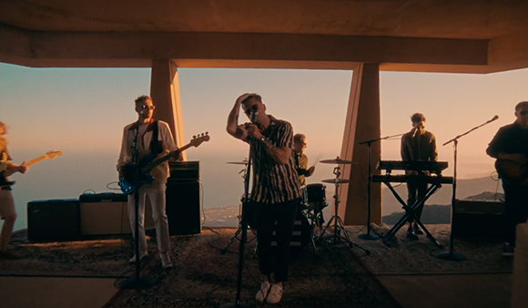 A band performs on a stage outdoors against a sunset sky.