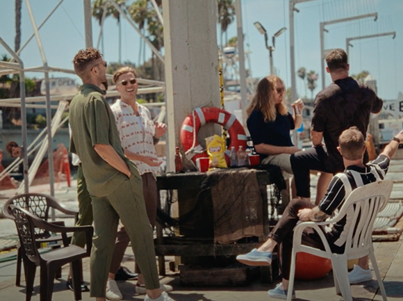 A group of young adults talking and sitting around a table near a pier on a sunny day.