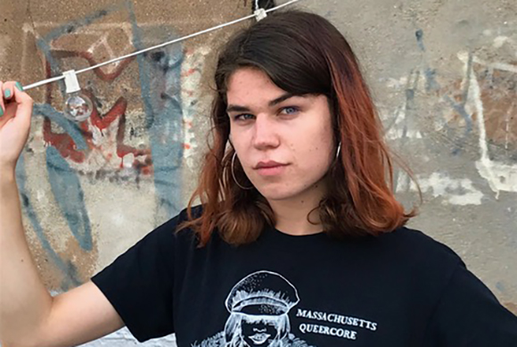 A person with reddish-brown hair wearing a black Massachusetts Queercore t-shirt in front of a graffiti wall.