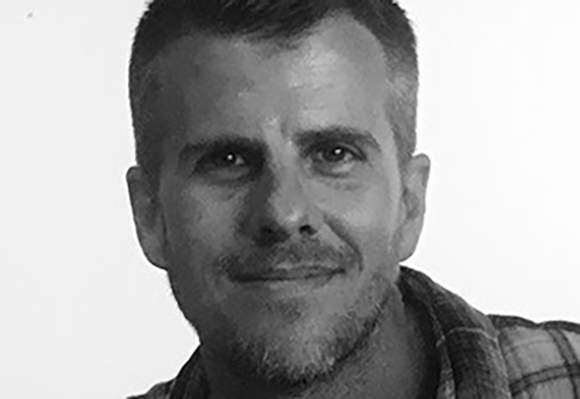 Black and white close-up portrait of a man with short hair and a light beard smiling.