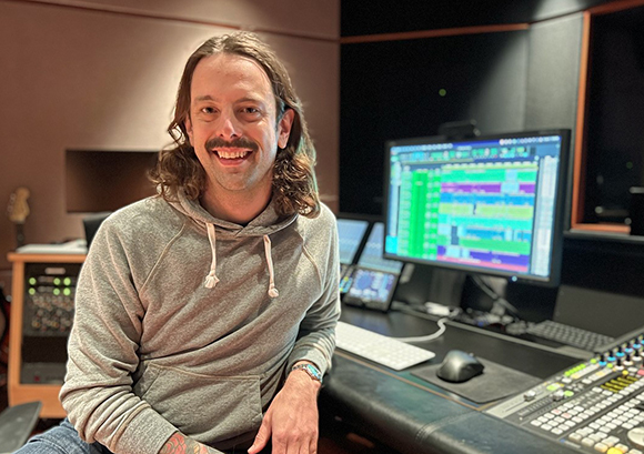 A man in a grey hoodie smiles in front of computer monitors and audio mixing equipment.