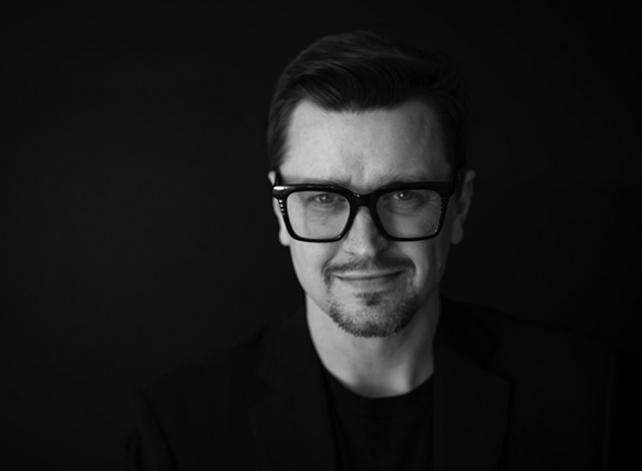Black and white portrait of a man with dark hair, glasses, and a goatee against a black background.