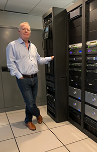 A man in a light blue shirt and jeans stands beside tall black server racks in a data center.