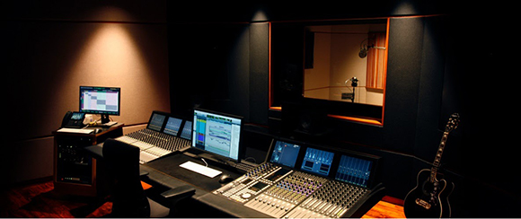 A dimly lit recording studio featuring a large mixing console, computer monitors, and an acoustic guitar.