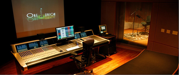 A large audio mixing console in a dark studio with a monitor and a view into a recording booth.