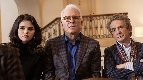 A young woman with dark hair and two older men sit at a table, looking toward the camera with serious expressions.