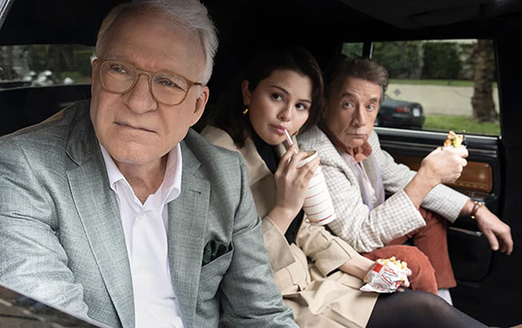 A man in a grey blazer, a woman with a drink, and a man eating popcorn sit in a car.