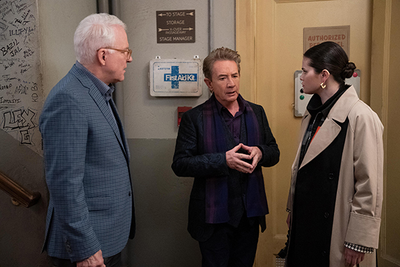 A man in a blue blazer, a man in a black jacket with a purple scarf, and a woman in a tan coat talk in a hallway.
